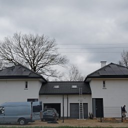 EcoEle - Nowoczesny, dwuczęściowy dom z panelami słonecznymi na dachu, widoczny z przodu, z samochodem i furgonetką zaparkowanymi przed garażem, drabina oparta o dach, pochmurne niebo w tle.