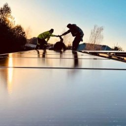 EcoEle - Instalacja paneli fotowoltaicznych na dachu w promieniach zachodzącego słońca, z widocznymi trzema monterami w odblaskowych kamizelkach podczas łączenia modułów.