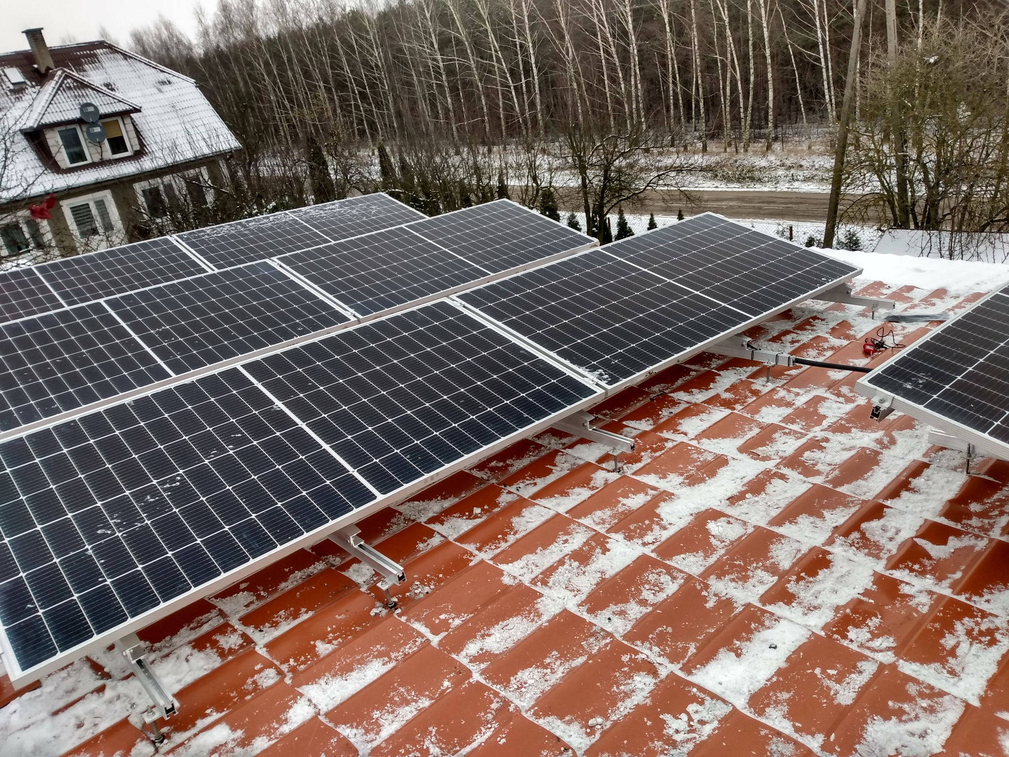 Panele słoneczne na pomarańczowym dachu pokrytym śniegiem, z widokiem na dom i las w tle.