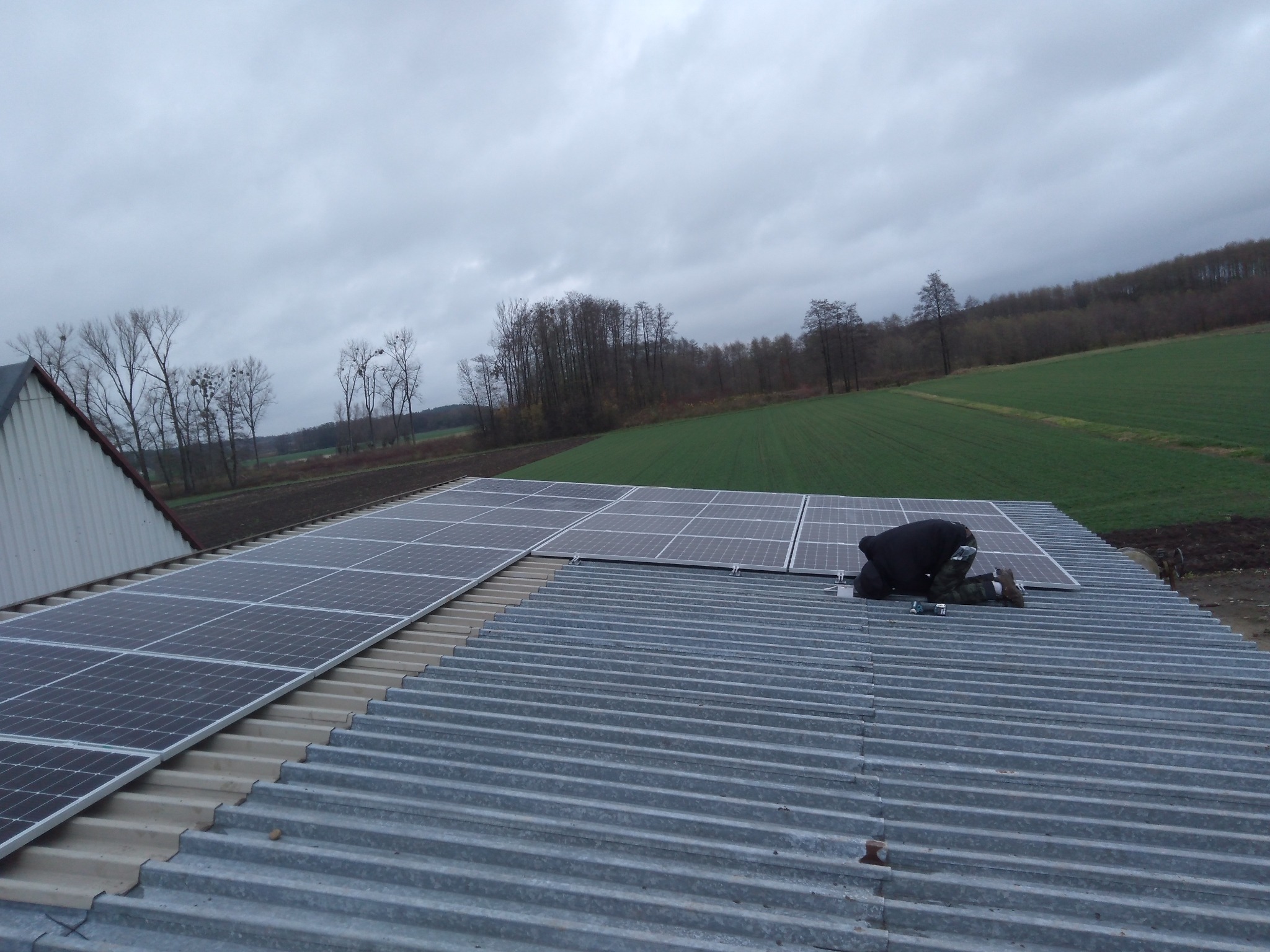 Montaż paneli słonecznych na blaszanym dachu, widok z góry na instalatora pracującego w pochmurny dzień, z polami i drzewami w tle.