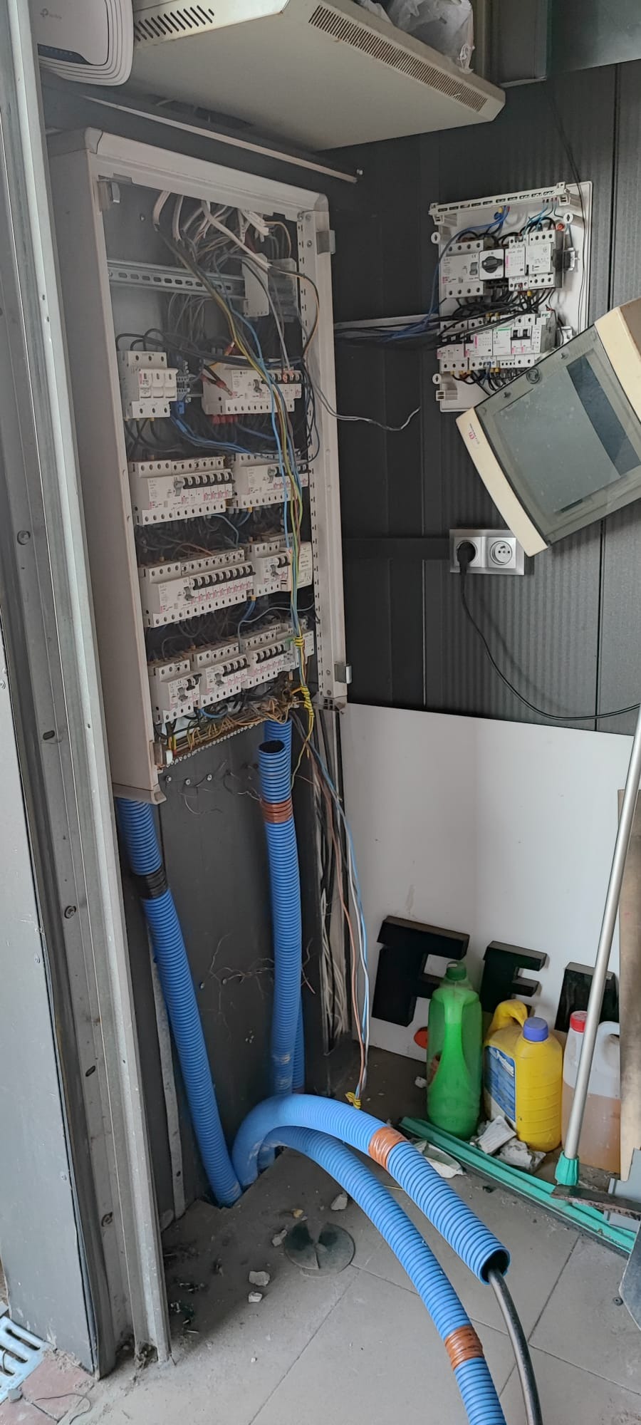 Otwarta szafa elektryczna z widocznymi bezpiecznikami, przewodami i niebieskimi rurami karbowanymi, z dodatkową skrzynką rozdzielczą na ścianie, w tle widoczne pojemniki z płynami i litery F i E.