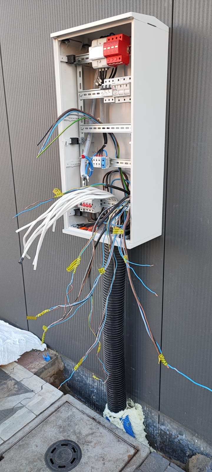 Biała skrzynka elektryczna z widocznymi zabezpieczeniami i przewodami, zamontowana na szarej ścianie z pionowymi żłobieniami. Przewody wychodzą z dołu skrzynki i są prowadzone w czarnej karbowanej...