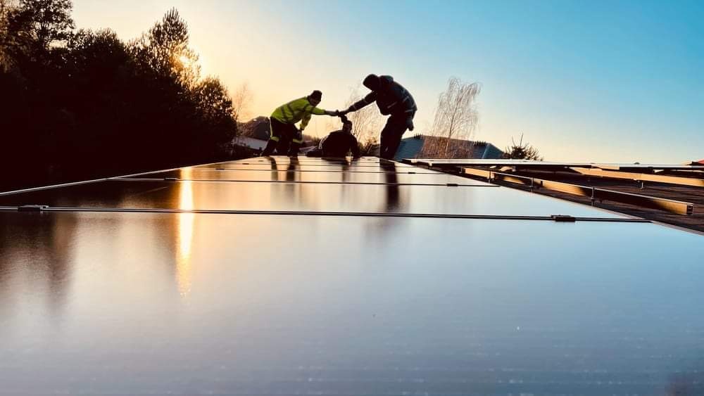 Instalacja paneli fotowoltaicznych na dachu w promieniach zachodzącego słońca, z widocznymi trzema monterami w odblaskowych kamizelkach podczas łączenia modułów.