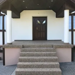 Wejście do domu z brązowymi drzwiami, kolumnami i schodami pokrytymi drobnym kamieniem. Podsufitka w kolorze ciemnego drewna.