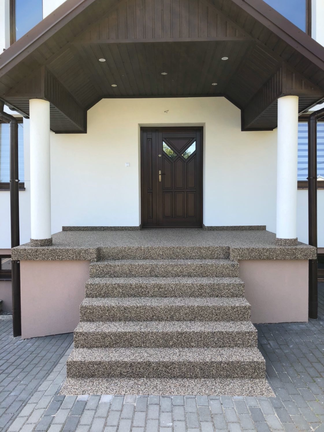 Wejście do domu z brązowymi drzwiami, kolumnami i schodami pokrytymi drobnym kamieniem. Podsufitka w kolorze ciemnego drewna.