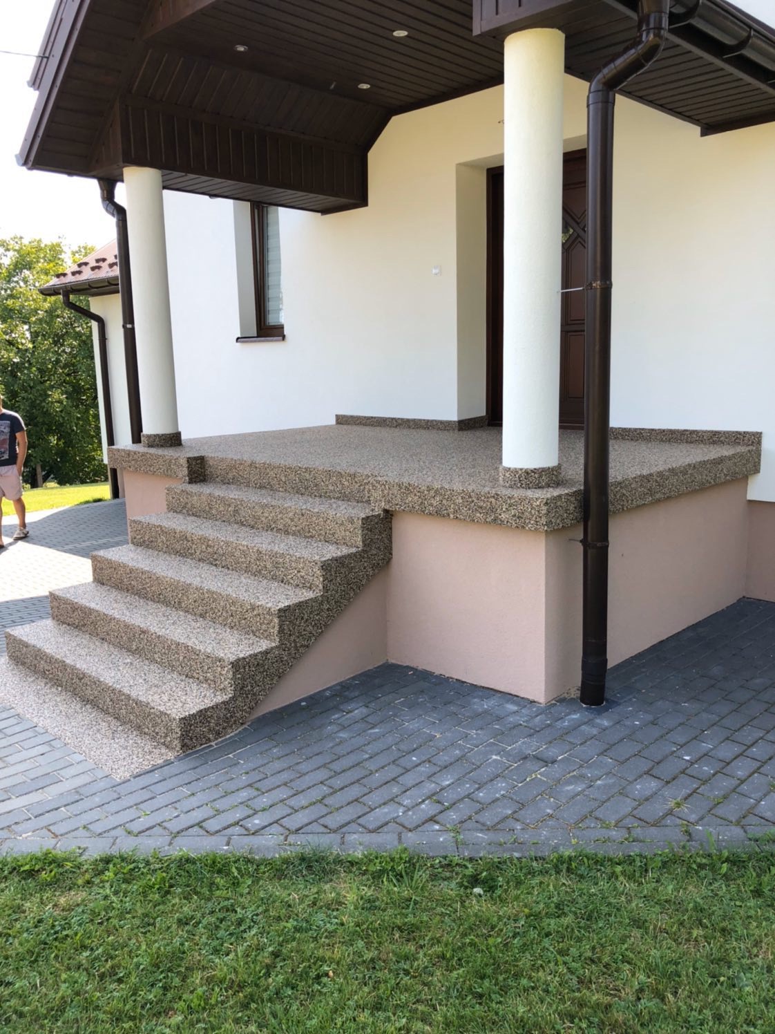 Schody wejściowe i podest pokryte beżową, kamyczkową nawierzchnią żywiczną, z widoczną teksturą i drobnymi detalami, prowadzące do domu z białymi ścianami i brązowymi elementami wykończeniowymi.