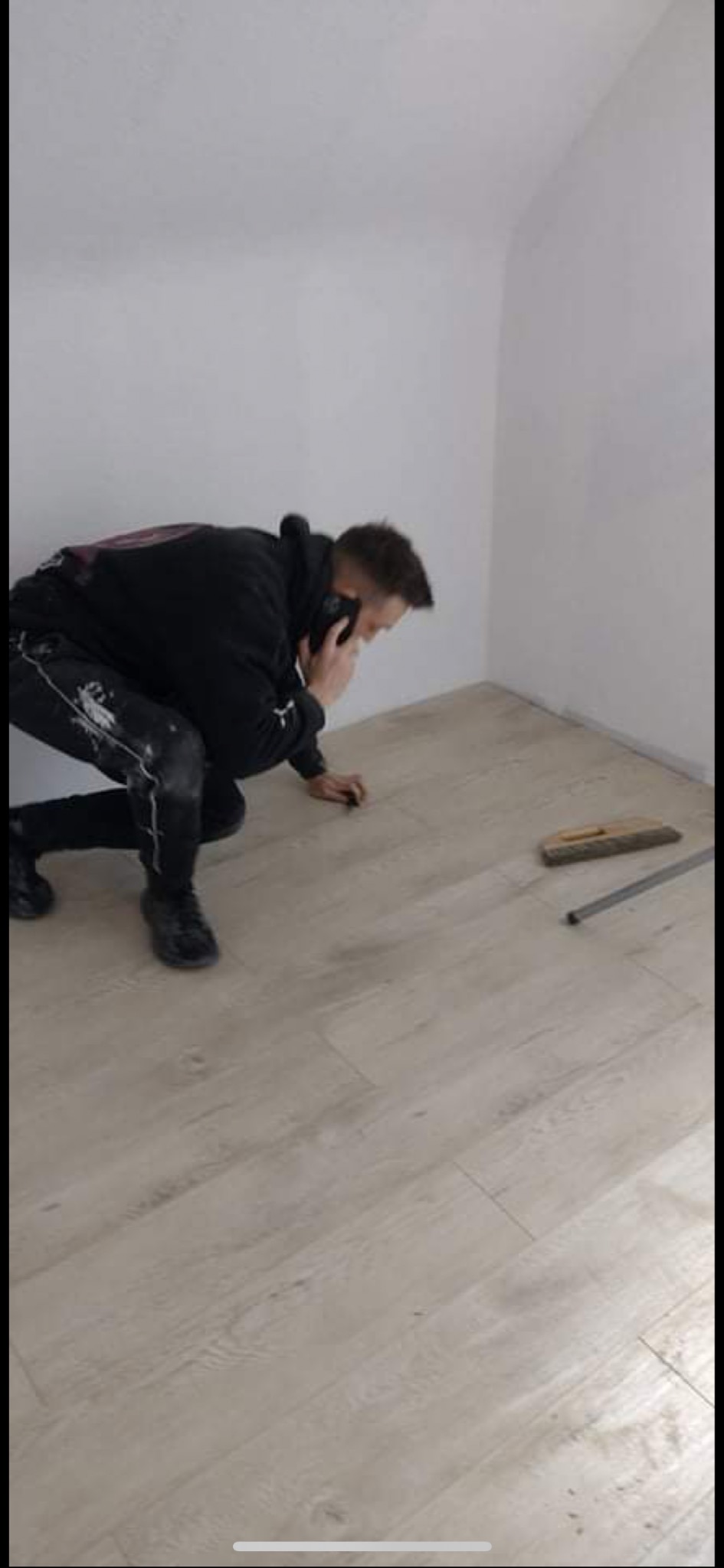 Pracownik w czarnej bluzie i spodniach z plamami klęczy na nowo położonej, jasnej podłodze laminowanej, rozmawiając przez telefon w rogu pokoju z białymi ścianami, obok leżą narzędzia.