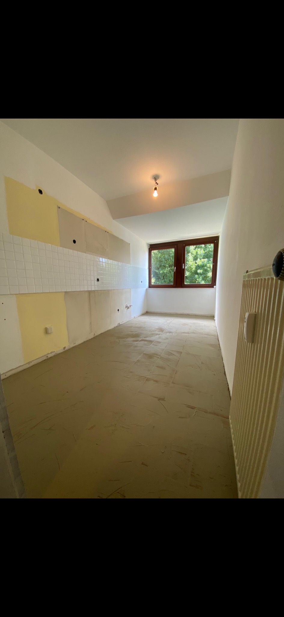 Wnętrze remontowanego pomieszczenia z przygotowanym podłożem pod wykładzinę, częściowo położone płytki na ścianie i widoczne ślady po malowaniu.