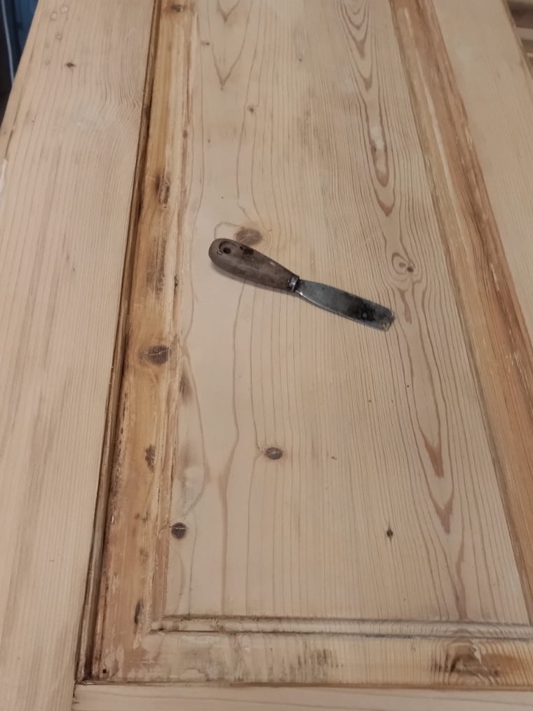 Fragment drewnianych drzwi z widocznymi śladami starej farby i szpachelką z drewnianą rączką, leżącą na powierzchni skrzydła drzwiowego.