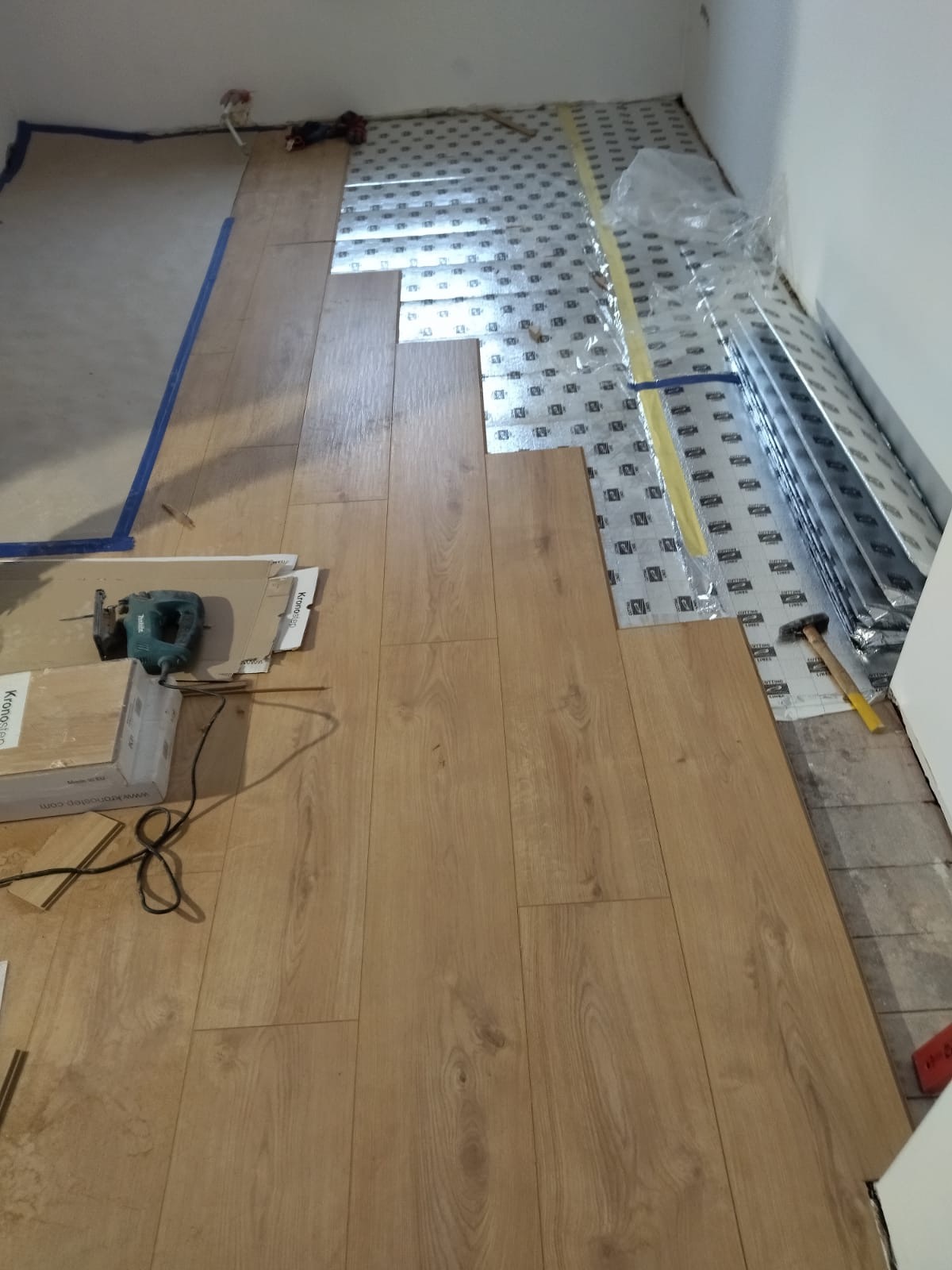 Układanie paneli podłogowych w trakcie pracy, widoczne podkłady izolacyjne, narzędzia (wyrzynarka, młotek) i opakowania paneli Kronostep.