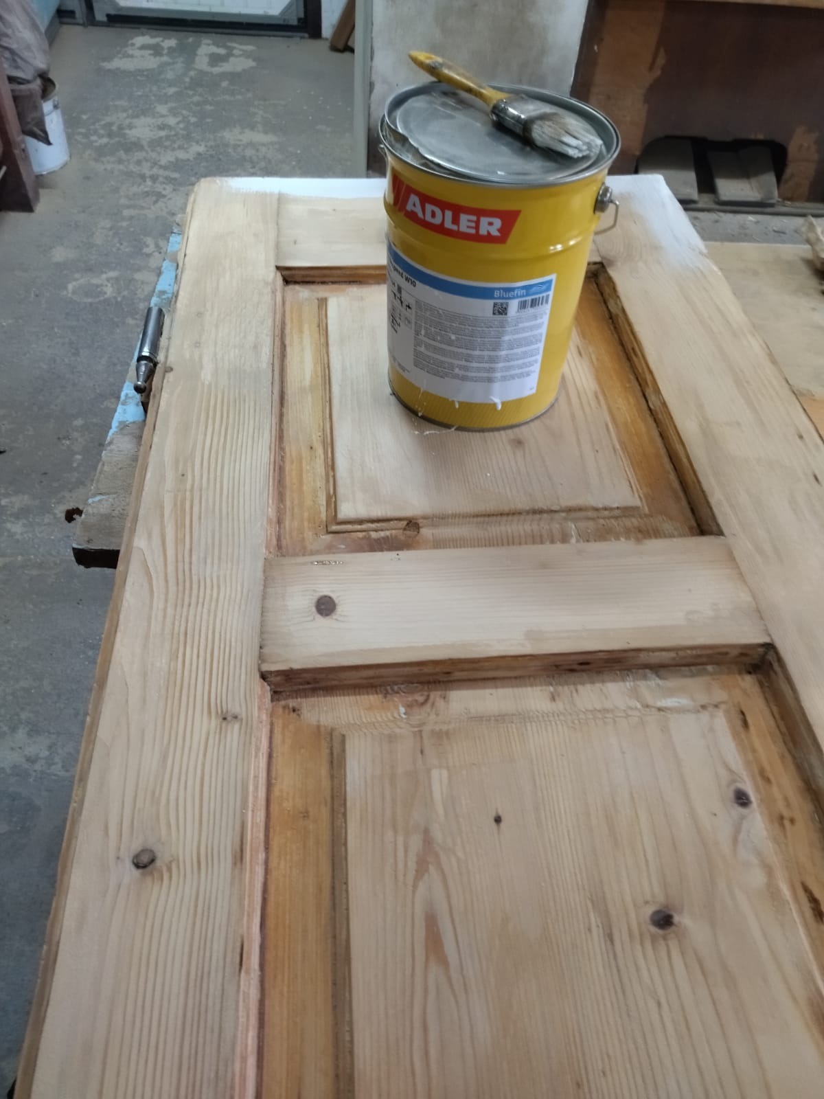 Drewniane drzwi z częściowo nałożoną białą farbą, obok puszka z farbą marki Adler i pędzel