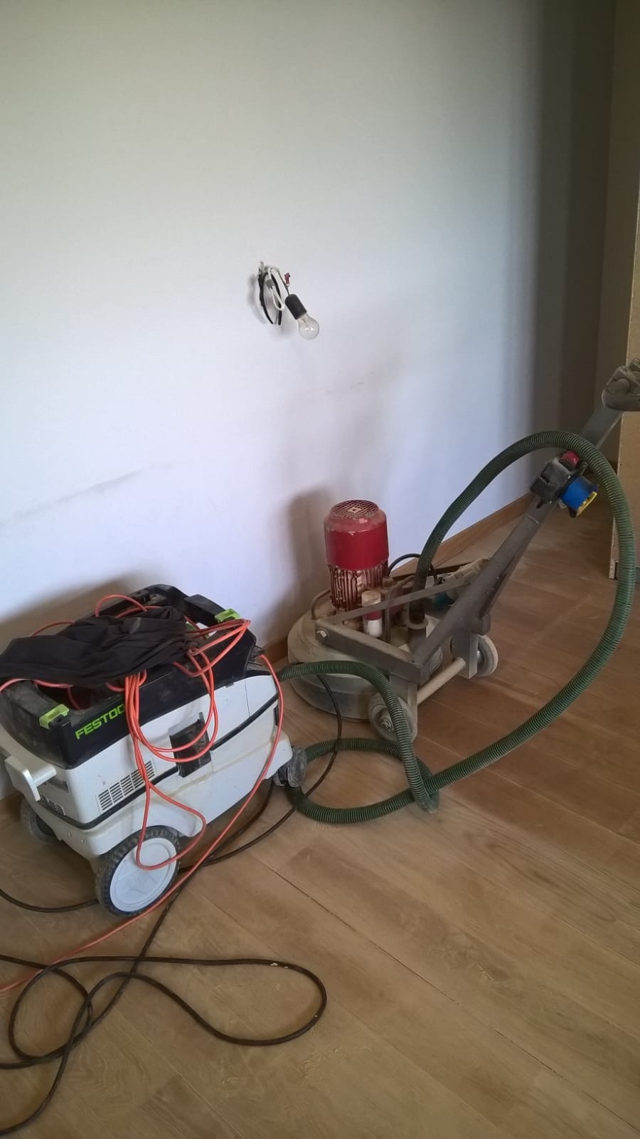 Parkieciarz używa szlifierki do podłóg Festool połączonej z odkurzaczem przemysłowym, widoczny kabel zasilający i niedokończona biała ściana z odsłoniętą żarówką.