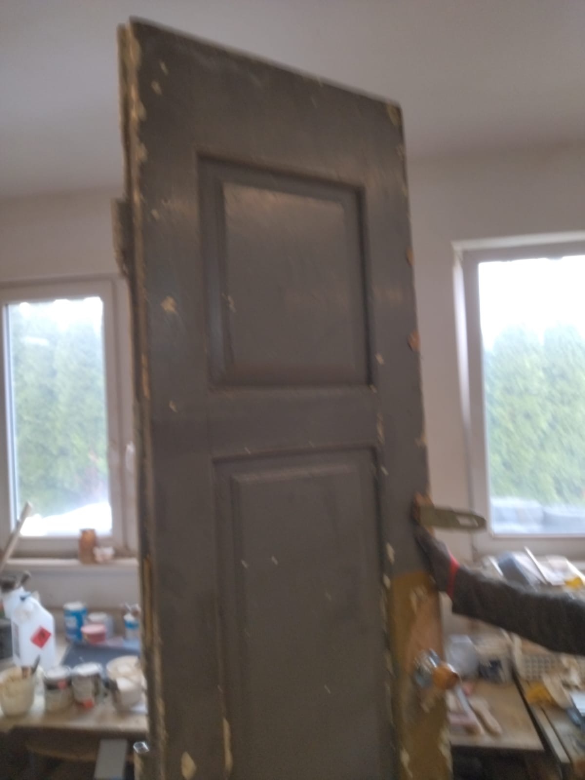 Stara, dwuskrzydłowa, szara, drewniana brama z widocznymi śladami zużycia i odchodzącą farbą, częściowo zasłaniająca widok na okno i zagracony stół warsztatowy z chemikaliami.