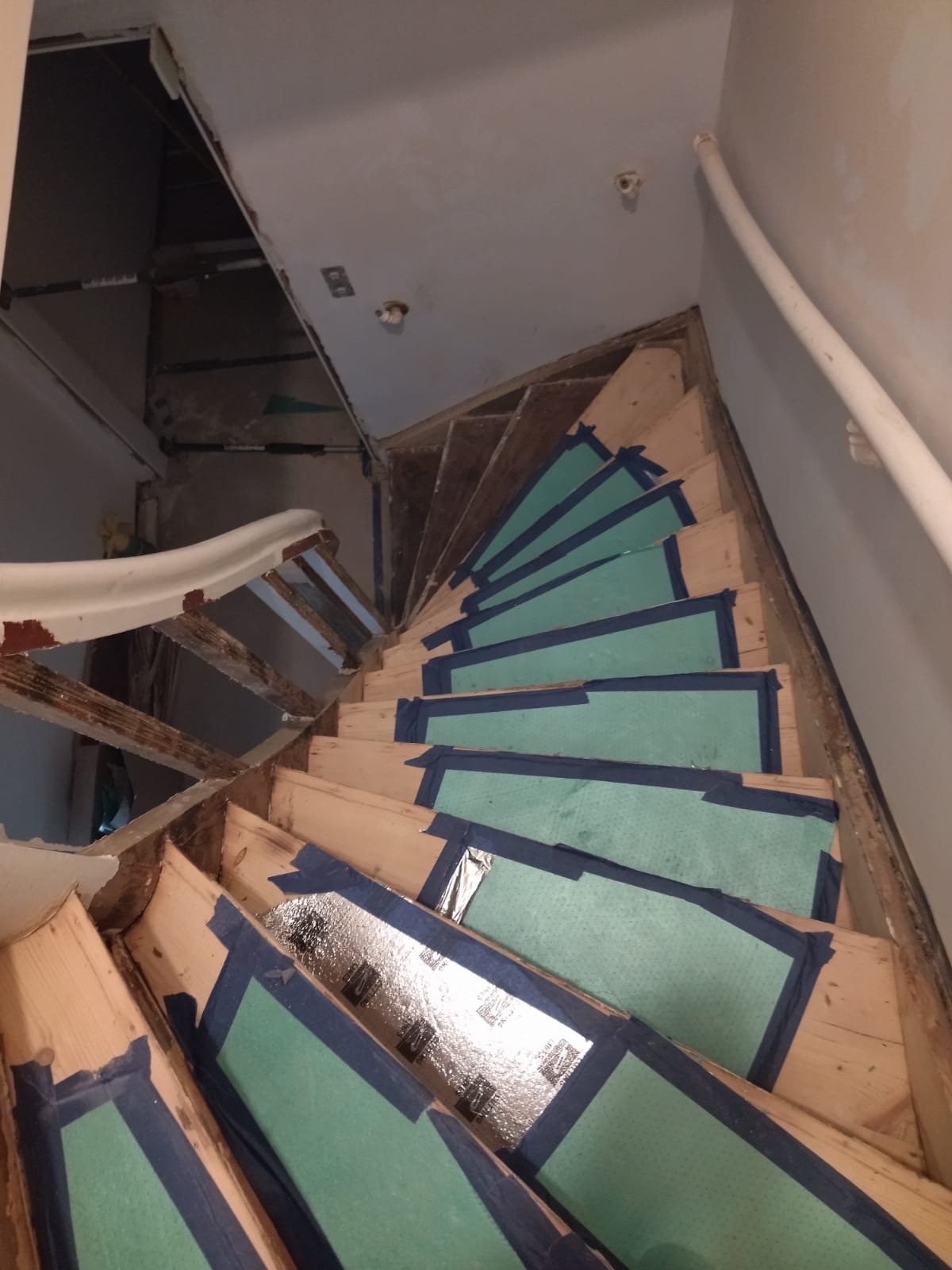 Schody drewniane w trakcie zabezpieczania folią i taśmą malarską przed remontem; widoczne niedokończone wykończenia ścian i balustrady.