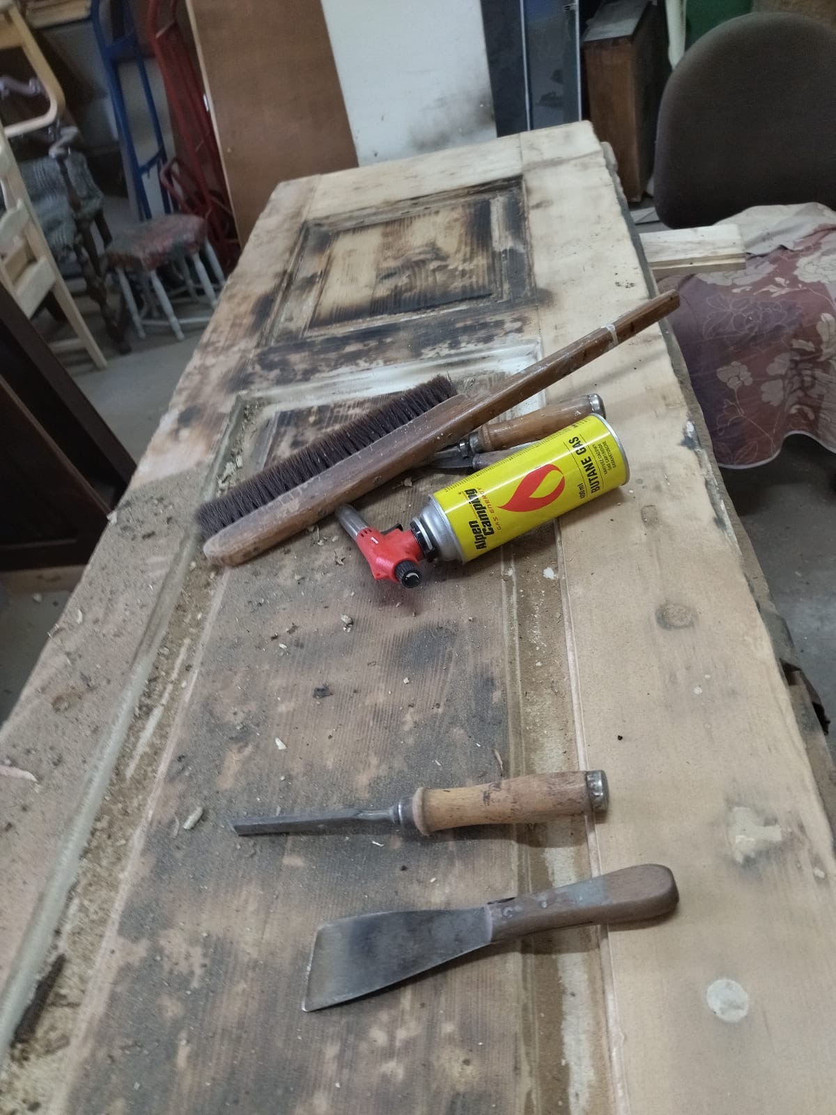 Stara, drewniana, brudna, drzwi poddawana renowacji, na której leżą narzędzia: szczotka, opalarka na gaz, dłuto i szpachelka.