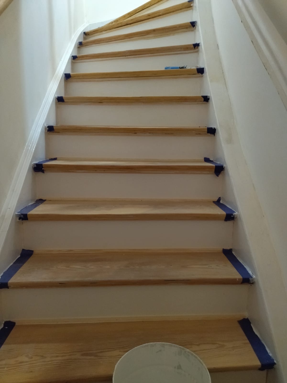 Schody drewniane zabezpieczone taśmą malarską, przygotowane do malowania ścian na biało, widoczny fragment wiadra z farbą.