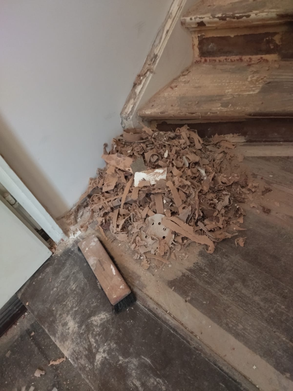 Stare drewniane schody w trakcie remontu, zeskrobane fragmenty farby i pył zebrane w kupkę, obok leży szczotka do zamiatania.