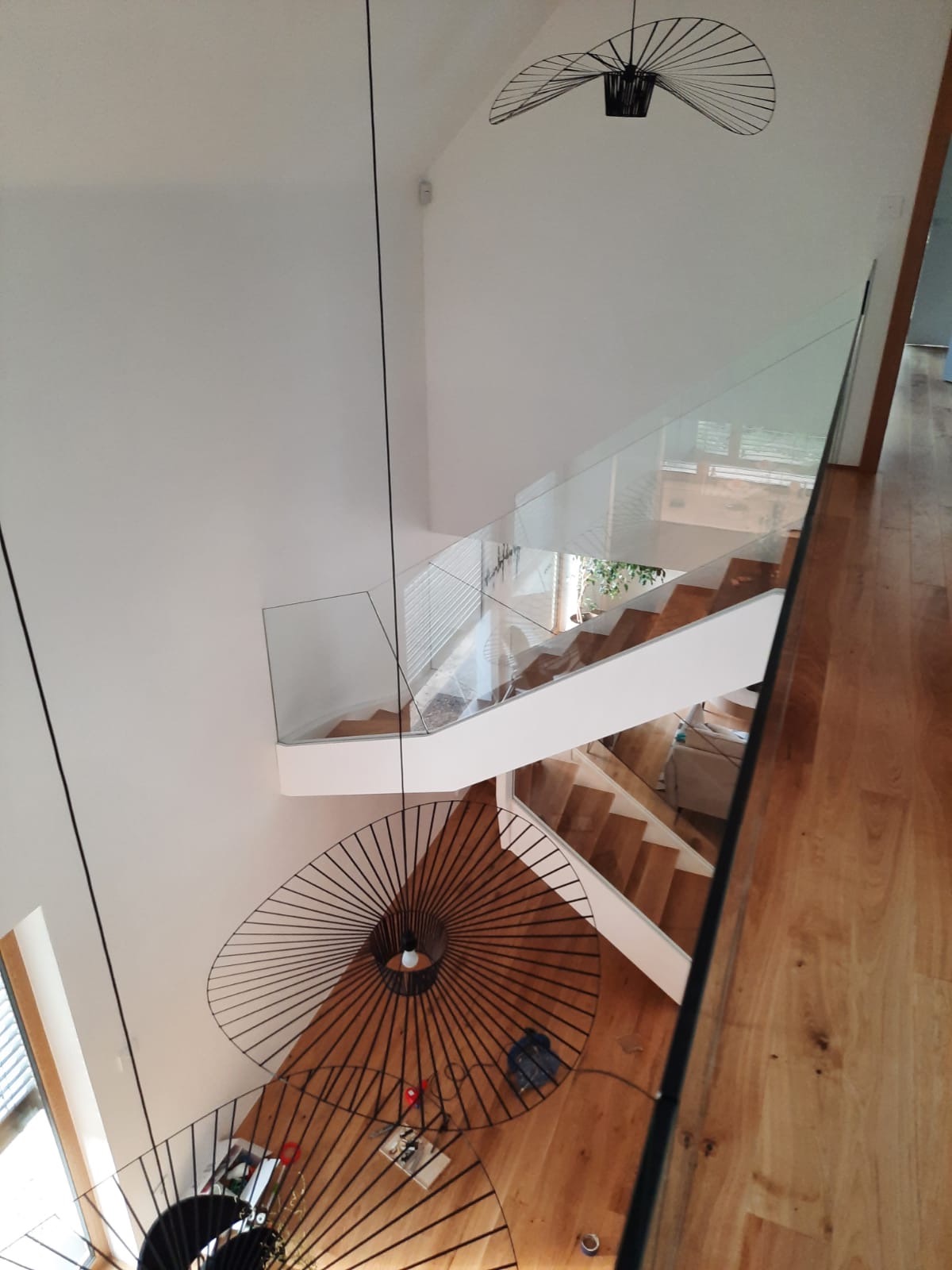 Wysoki hol z drewnianymi schodami i szklanymi balustradami, widok z góry. Dwie nowoczesne lampy wiszące z czarnymi, metalowymi kloszami. Świeżo pomalowane białe ściany i drewniana podłoga.