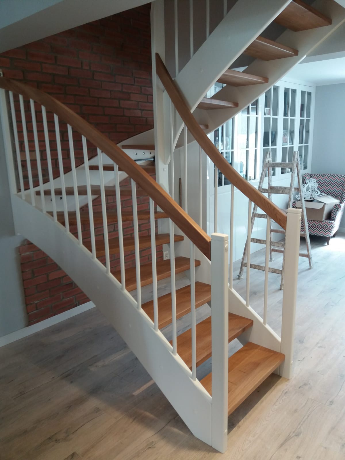 Nowoczesne, drewniane schody z białymi balustradami na tle ściany z czerwonej cegły i jasnej podłogi, z drabina i fotelem w tle.