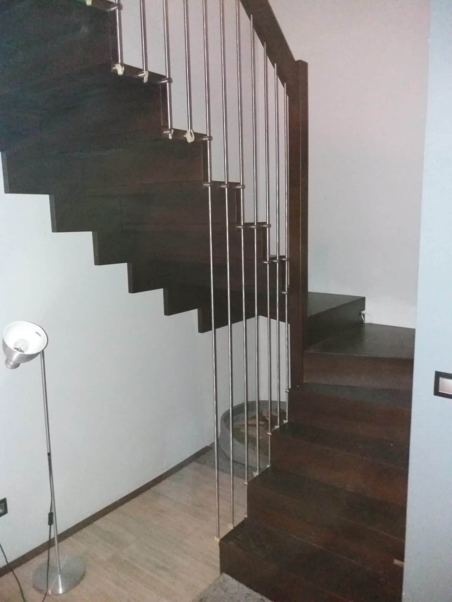 Drewniane schody z metalową balustradą w nowoczesnym wnętrzu, oświetlone lampą podłogową z regulowanym kloszem.