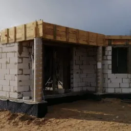 Budowa domu jednorodzinnego w trakcie wznoszenia ścian z bloczków, widoczne szalunki drewniane i zabezpieczenie przeciwwilgociowe fundamentów, w tle drugi budynek i samochód.