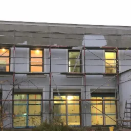 Ocieplanie budynku w Poznaniu, widoczne rusztowania i częściowo otynkowana elewacja, okna zabezpieczone folią, ciepłe światło w oknach.