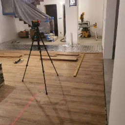 Wnętrze w trakcie remontu: układanie paneli podłogowych z użyciem poziomicy laserowej, zabezpieczone schody folią malarską, widoczna piła stołowa w tle.