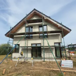 Dom jednorodzinny w trakcie prac elewacyjnych, widoczne rusztowania, okna z brązowymi ramami oraz piła do cięcia drewna na statywie.