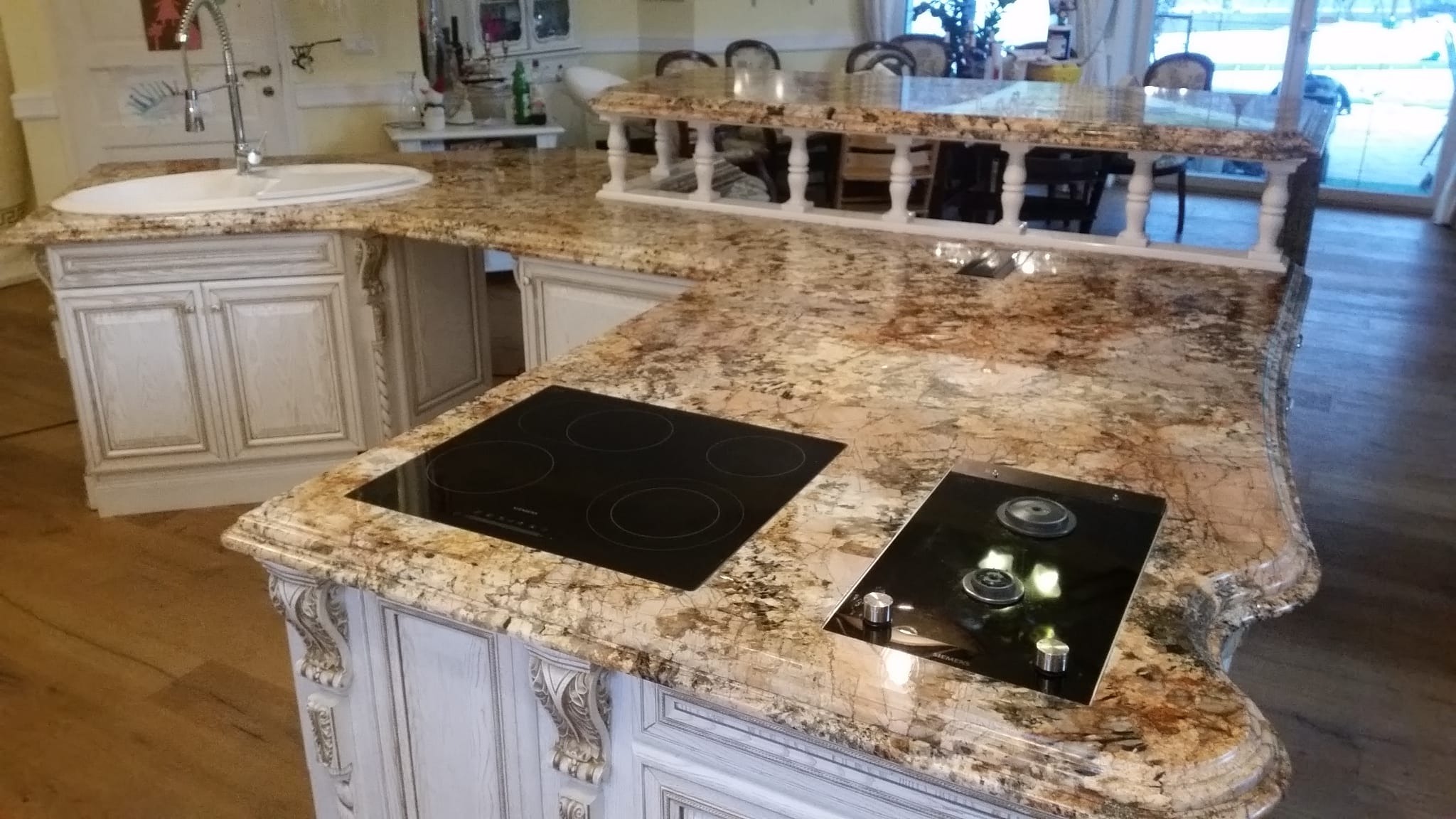 Kuchnia w stylu klasycznym z blatem z kamienia naturalnego, wbudowaną płytą indukcyjną i zlewozmywakiem, widoczne zdobione szafki.