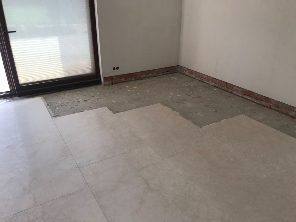 Układanie jasnych, marmurkowych płytek podłogowych w pokoju, widoczny fragment przygotowanego podłoża i ceglany cokół wzdłuż ściany.