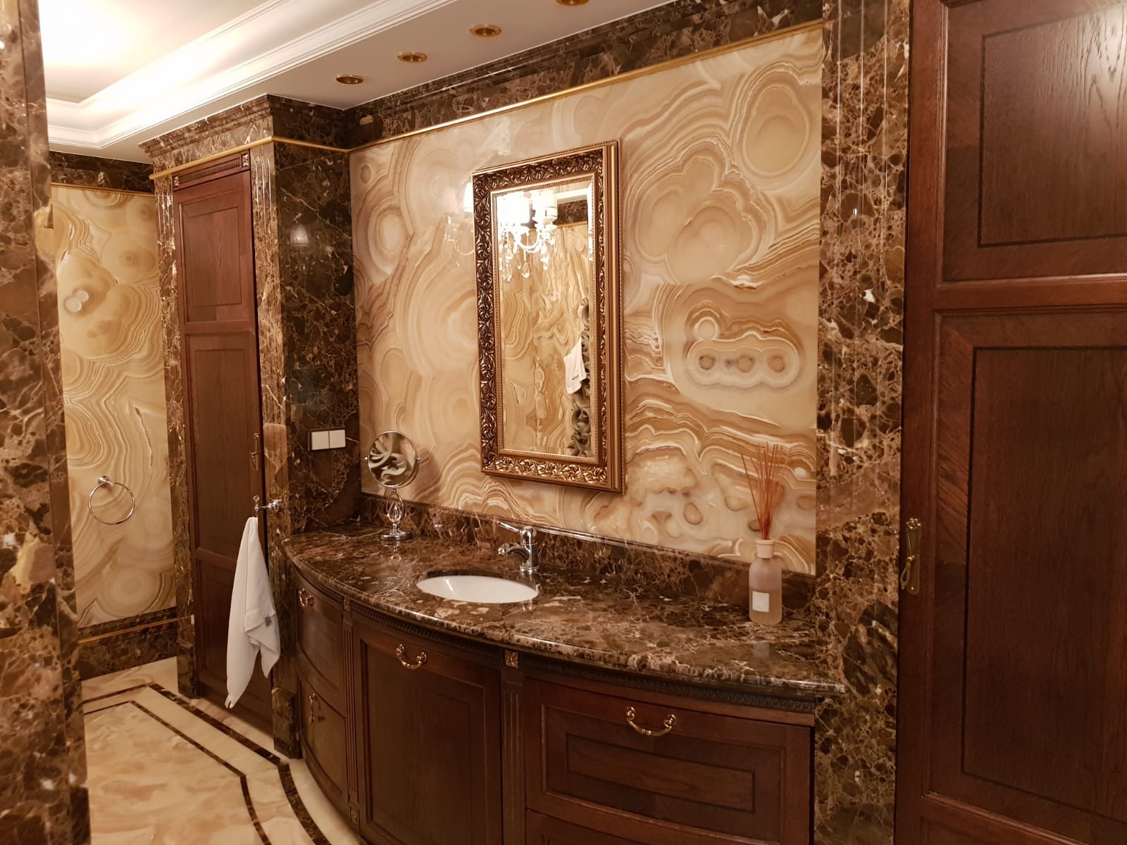 Elegancka łazienka z marmurowymi ścianami i blatem, ciemne drewniane szafki z ozdobnymi uchwytami, złota rama lustra i detale wykończeniowe, symetryczny układ elementów.