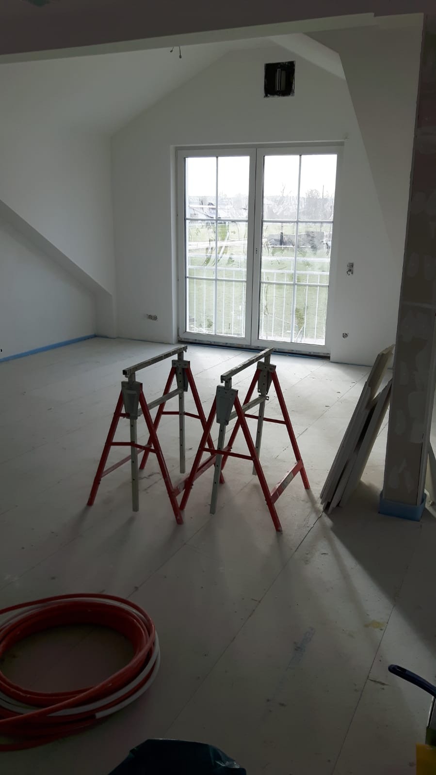 Poddasze w trakcie prac wykończeniowych: białe ściany, okno balkonowe, czerwone kozły malarskie, zwój rur grzewczych na podłodze, przygotowane płyty gipsowe przy ścianie.