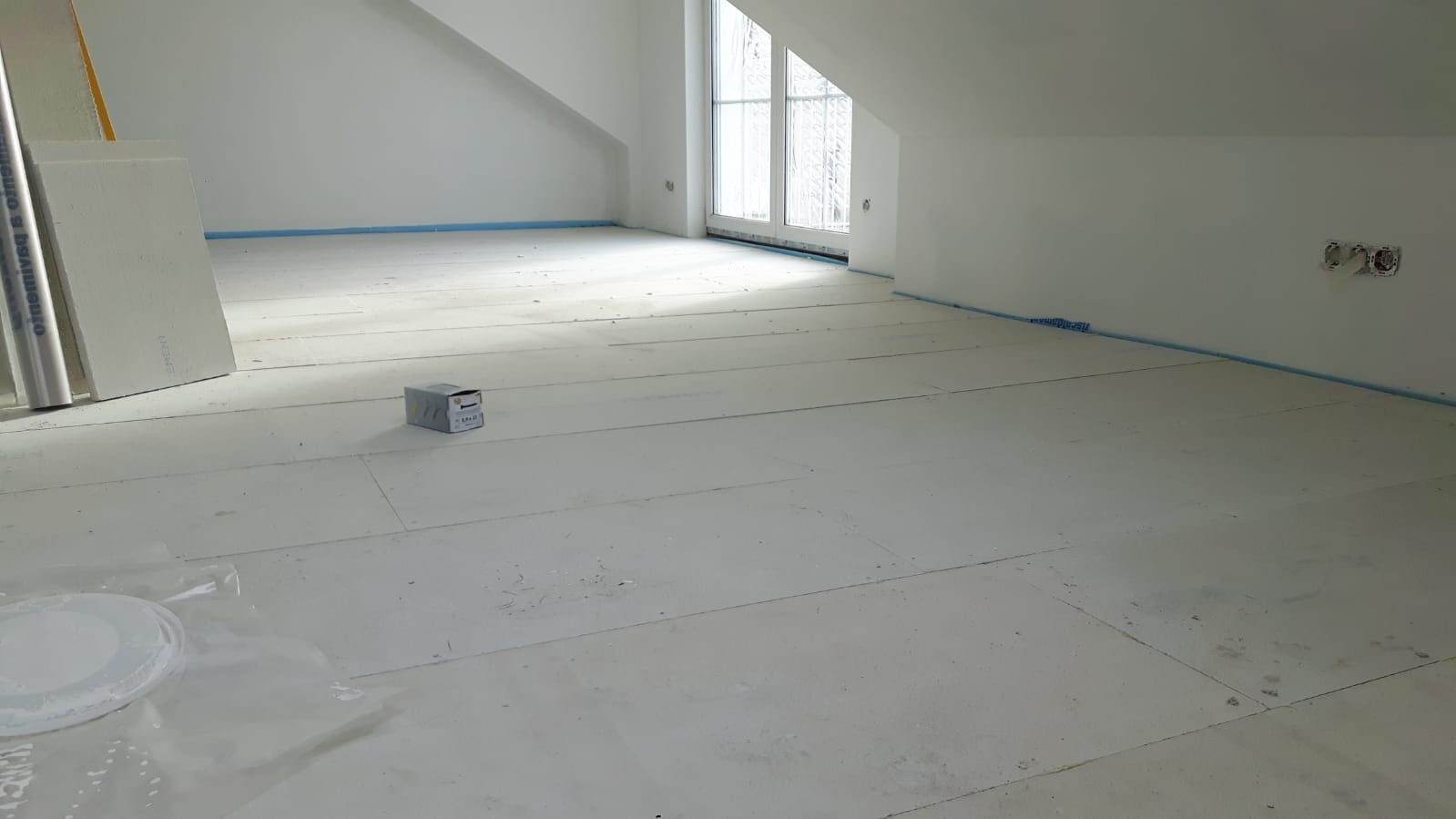 Poddasze w trakcie izolacji podłogi, widoczne płyty styropianowe, okno dachowe, zabezpieczona listwa przypodłogowa i puszki elektryczne.