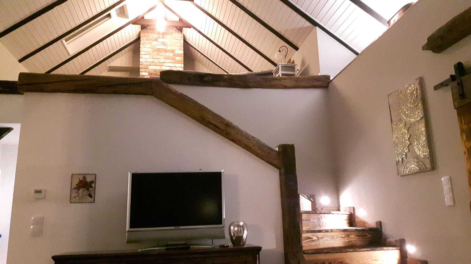 Wnętrze domu z drewnianymi belkami stropowymi, ceglanym kominem, drewnianymi schodami z podświetleniem, telewizorem i dekoracjami na ścianach.