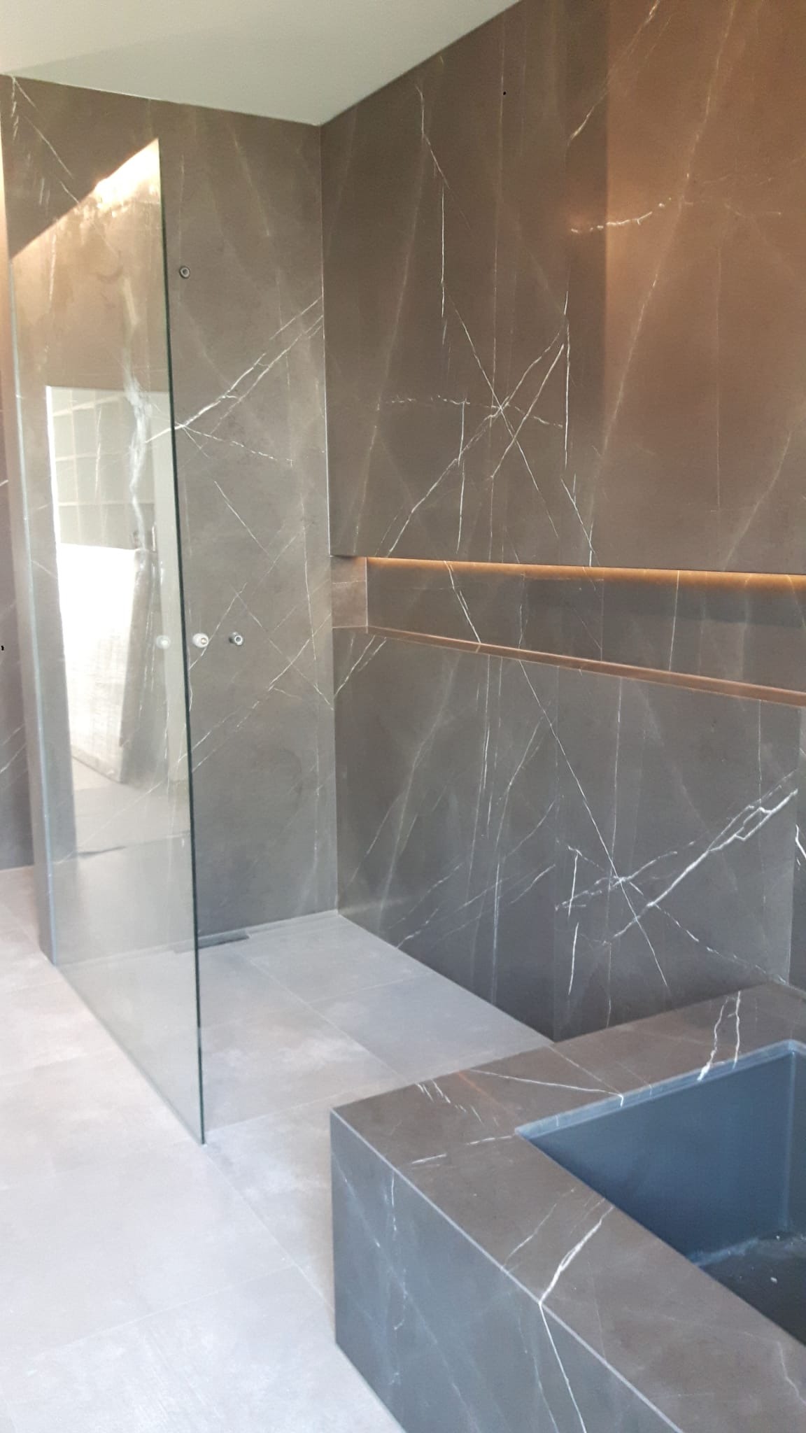 Nowoczesna łazienka z szarymi płytkami imitującymi marmur, kabina prysznicowa ze szkła hartowanego oraz zabudowana wanna, oświetlenie LED w półce.