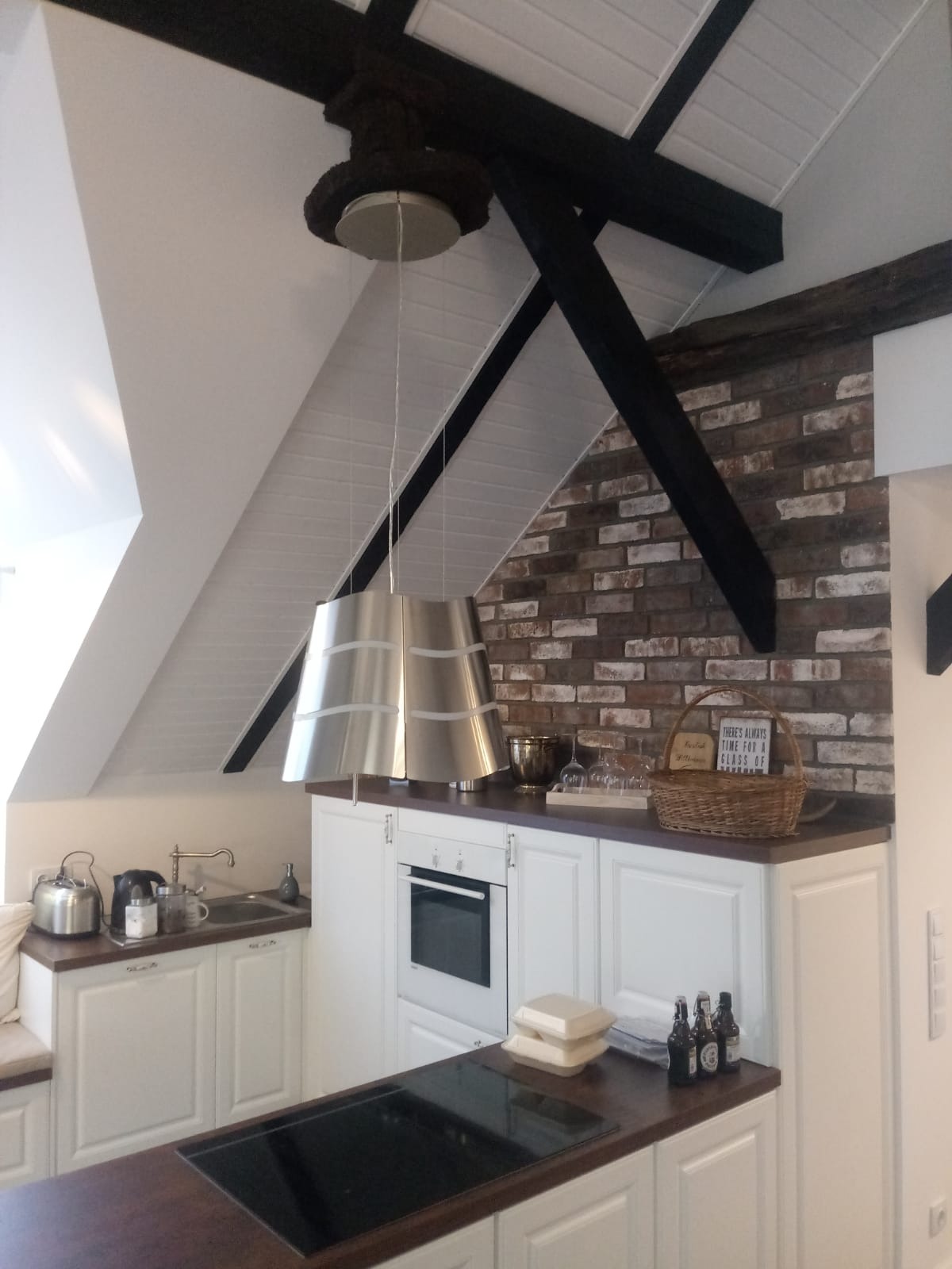 Kuchnia w stylu rustykalnym z białymi szafkami, drewnianym blatem i ceglaną ścianą, oświetlona metalowymi lampami wiszącymi, widoczne elementy dekoracyjne jak koszyk i butelki.