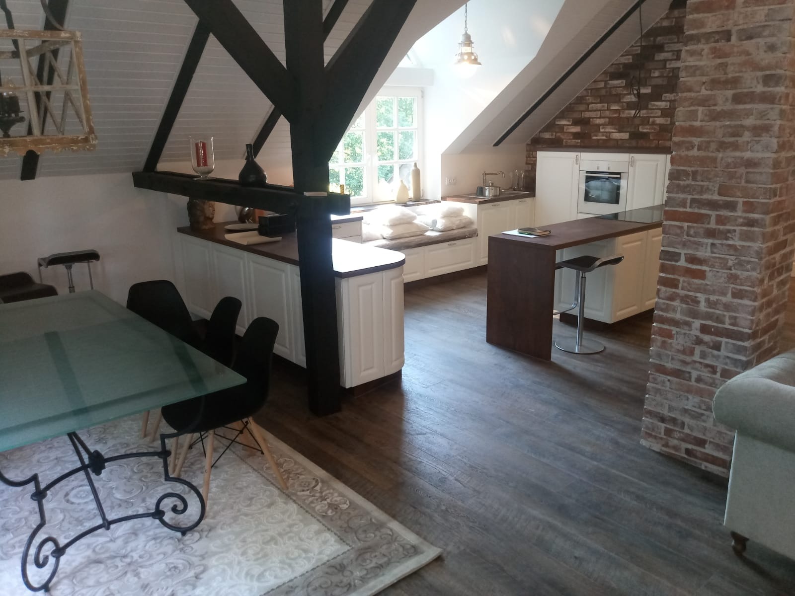 Kuchnia w stylu rustykalnym na poddaszu z drewnianymi belkami stropowymi, ceglaną ścianą i wyspą kuchenną z hokerem, oświetlona wiszącą lampą.