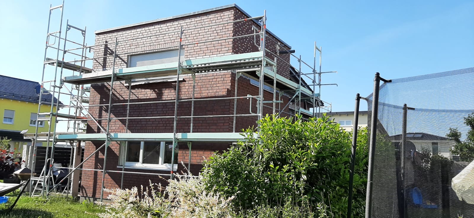 Dwupiętrowy dom z czerwonej cegły w trakcie prac z użyciem rusztowania, widoczny fragment ogrodu i trampoliny.