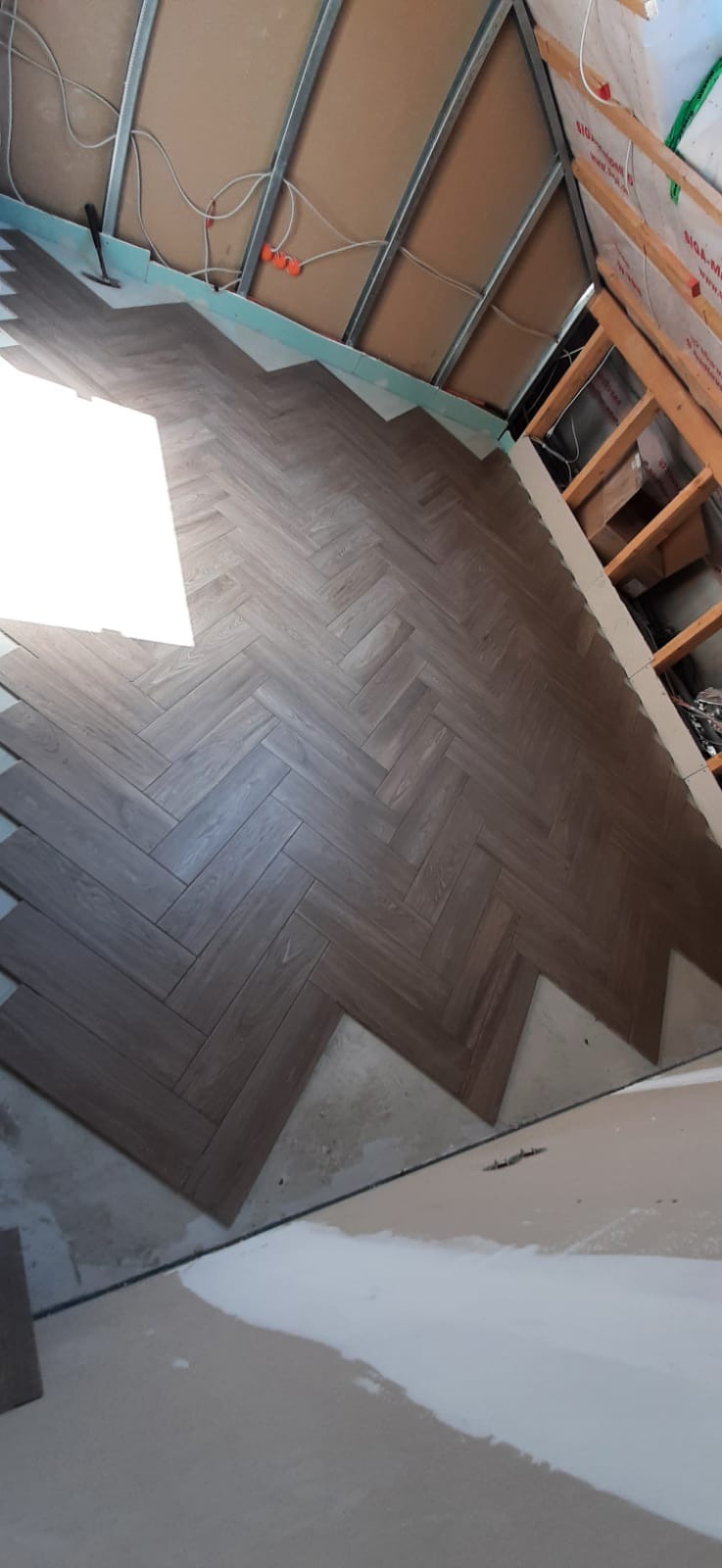 Układanie podłogi z płytek imitujących drewno w jodełkę, widoczne niedokończone krawędzie i elementy konstrukcyjne sufitu podwieszanego.