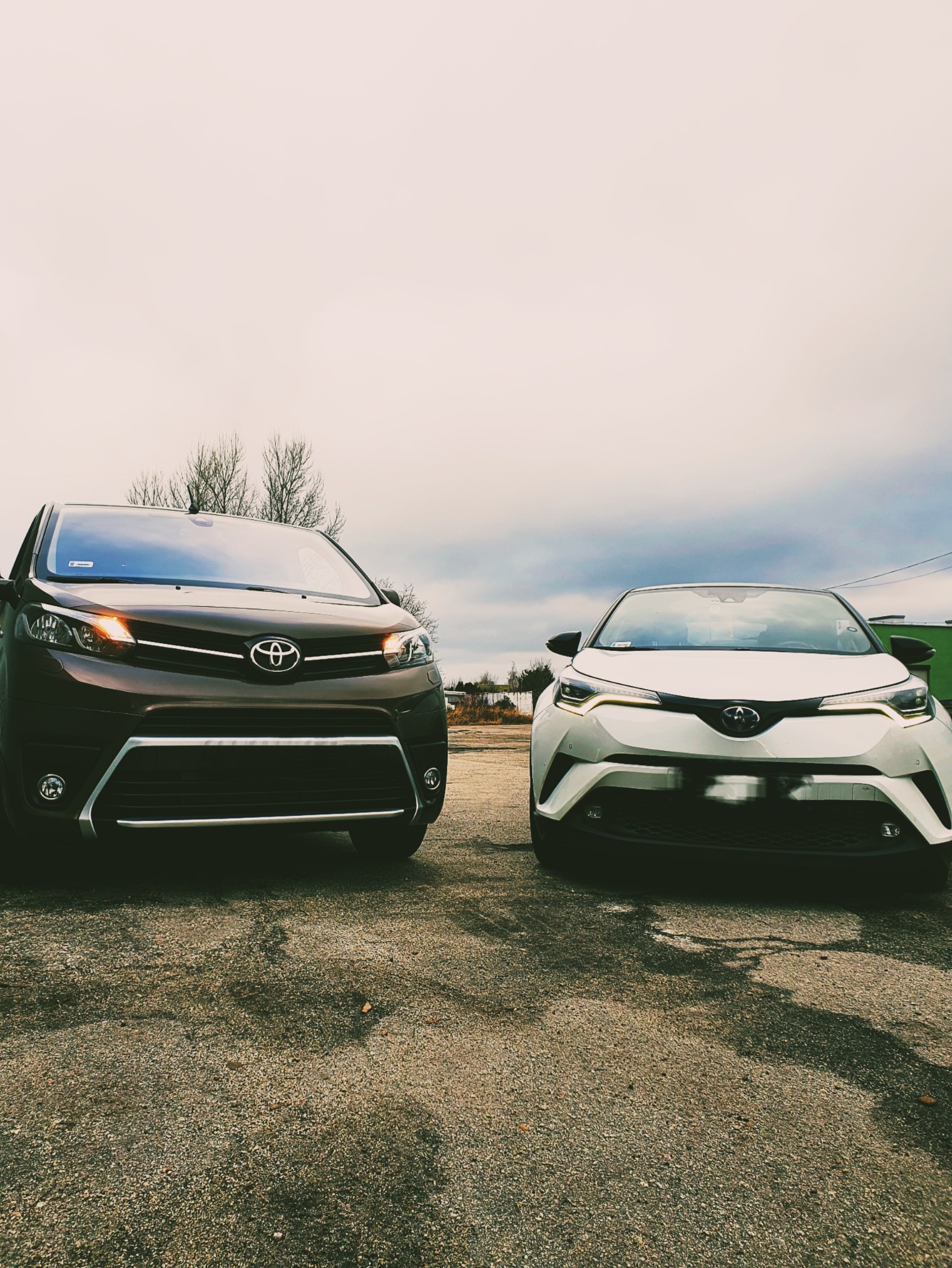 Dwa samochody osobowe marki Toyota, brązowy i biały, stojące obok siebie na szarym, żwirowym podłożu, z pochmurnym niebem w tle.