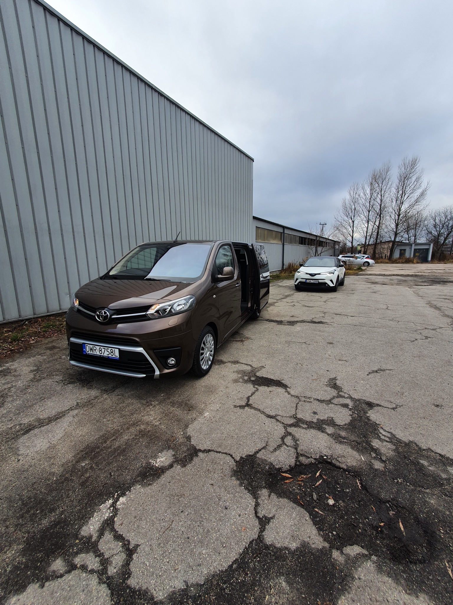 Brązowy van Toyota z otwartymi drzwiami przesuwnymi na tle szarego budynku przemysłowego i popękanego asfaltu, w tle zaparkowany biały samochód osobowy.