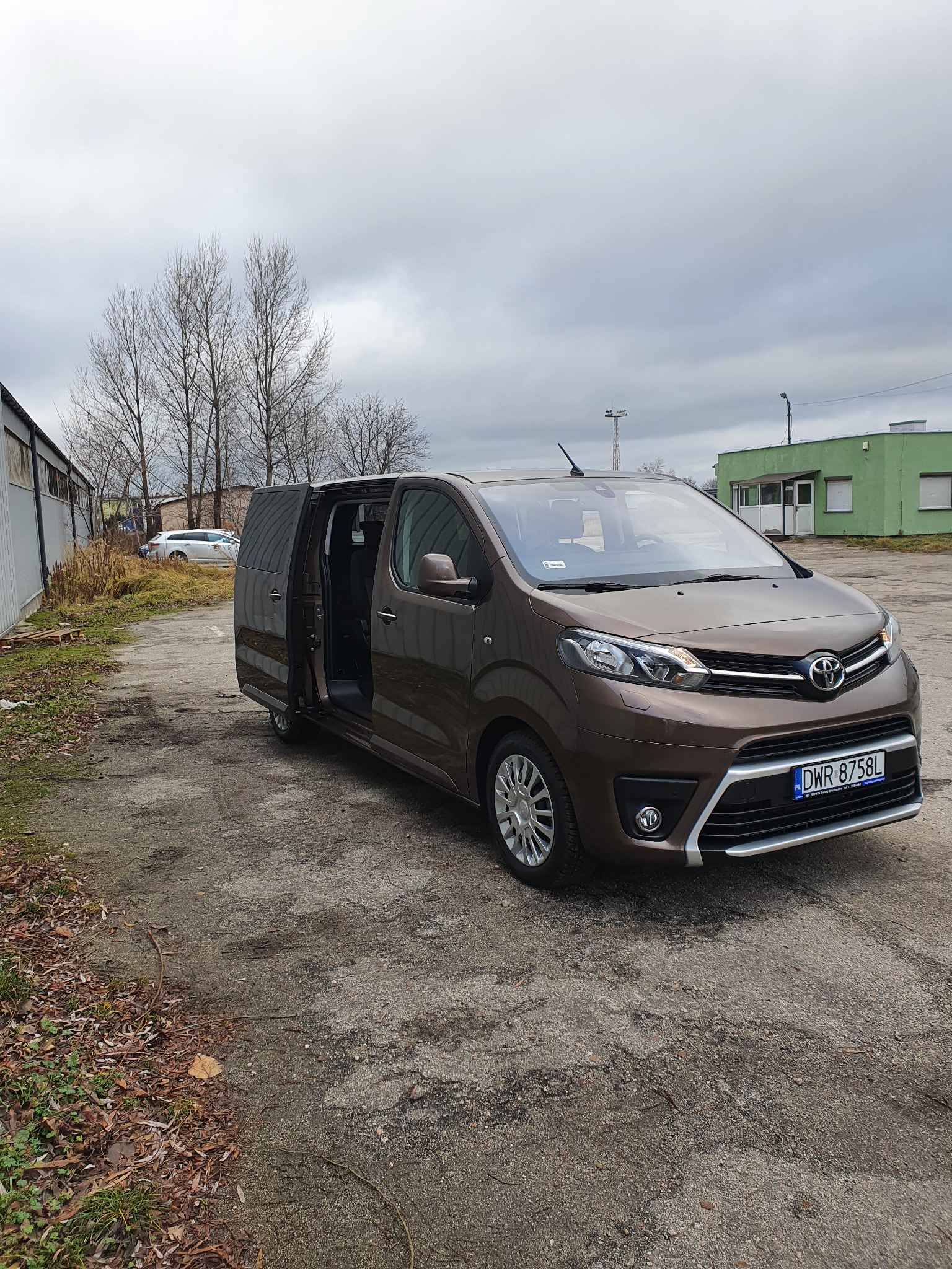 Brązowy van Toyota Proace Verso z otwartymi drzwiami przesuwnymi, zaparkowany na szarym placu, widoczne budynki w tle i pochmurne niebo.
