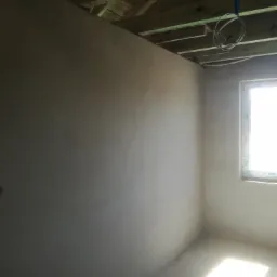 Świeżo otynkowane ściany w surowym wnętrzu z drewnianym stropem i widocznymi kablami elektrycznymi, doświetlone przez okno.