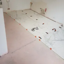 Układanie wielkoformatowych płytek imitujących marmur na podłodze, widoczne przyssawki do przenoszenia płytek i pomarańczowe klipsy poziomujące, surowe ściany z wystającymi przewodami elektrycznymi.