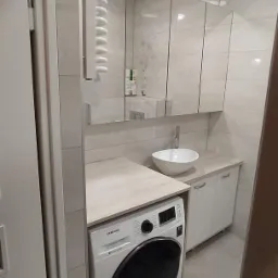 Nowoczesna łazienka z pralką zabudowaną pod blatem z umywalką nablatową, nad nią szafka lustrzana z białymi frontami.