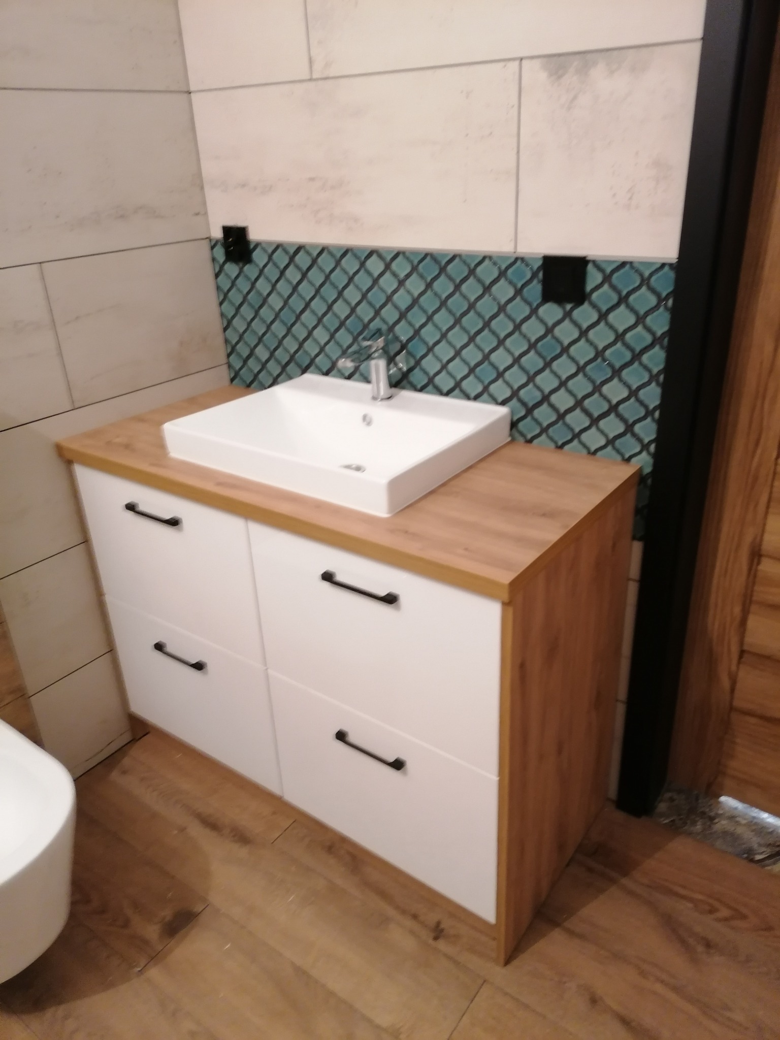 Nowoczesna łazienka z białą szafką pod umywalkę z drewnianym blatem, turkusowa mozaika na ścianie i drewniana podłoga.