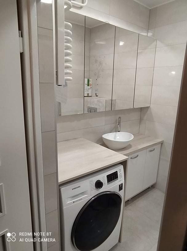 Nowoczesna łazienka z pralką zabudowaną pod blatem z umywalką nablatową, nad nią szafka lustrzana z białymi frontami.