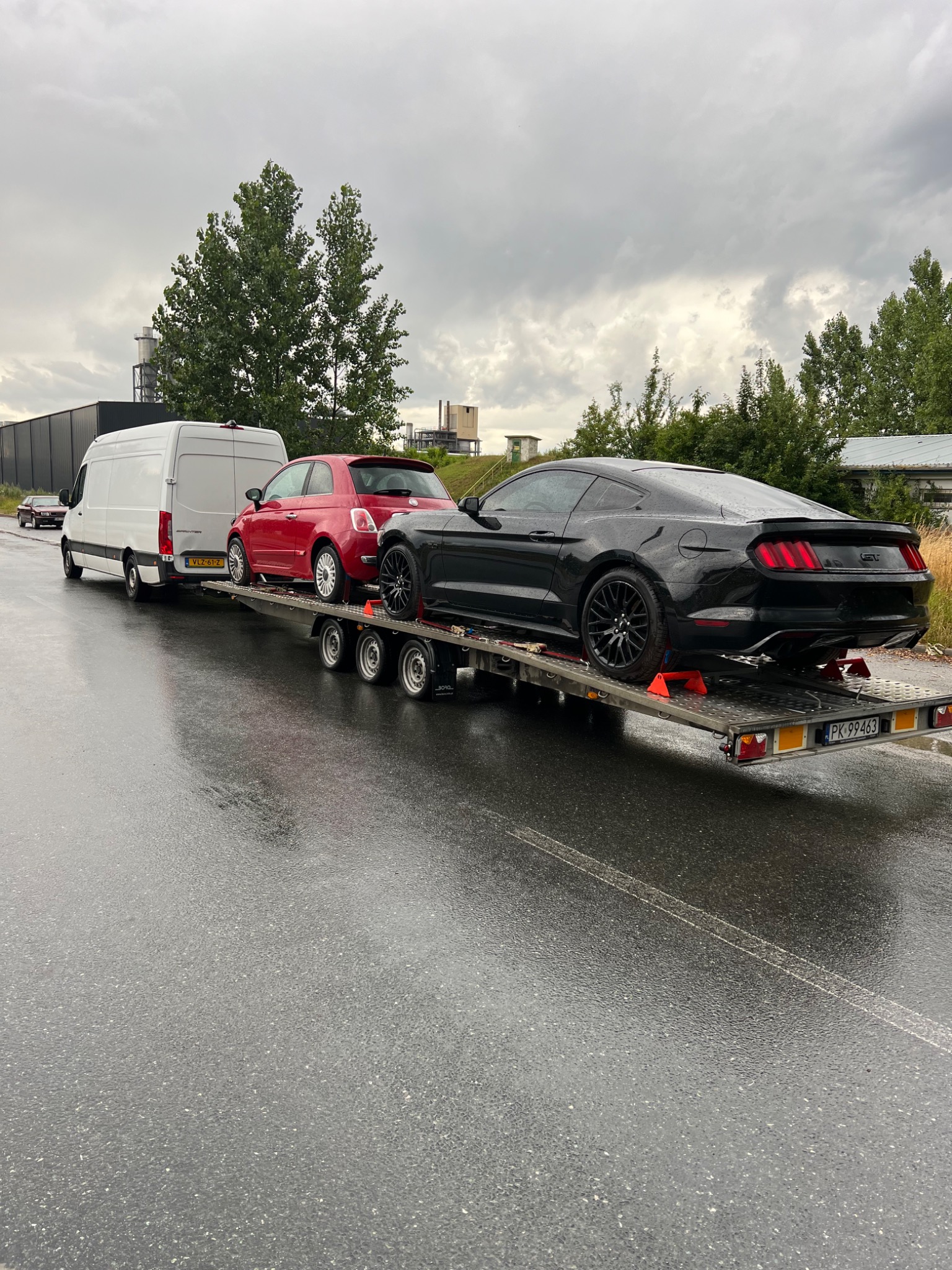 Laweta z trzema samochodami: białym vanem, czerwonym Fiatem 500 i czarnym Fordem Mustangiem, na mokrej nawierzchni drogi w pochmurny dzień. Widoczny budynek przemysłowy w tle.