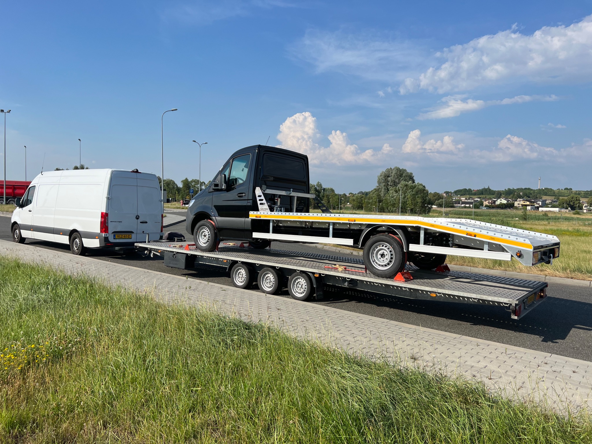 Laweta z czarnym samochodem na pokładzie, ciągnięta przez biały bus na tle letniego krajobrazu z błękitnym niebem i chmurami.