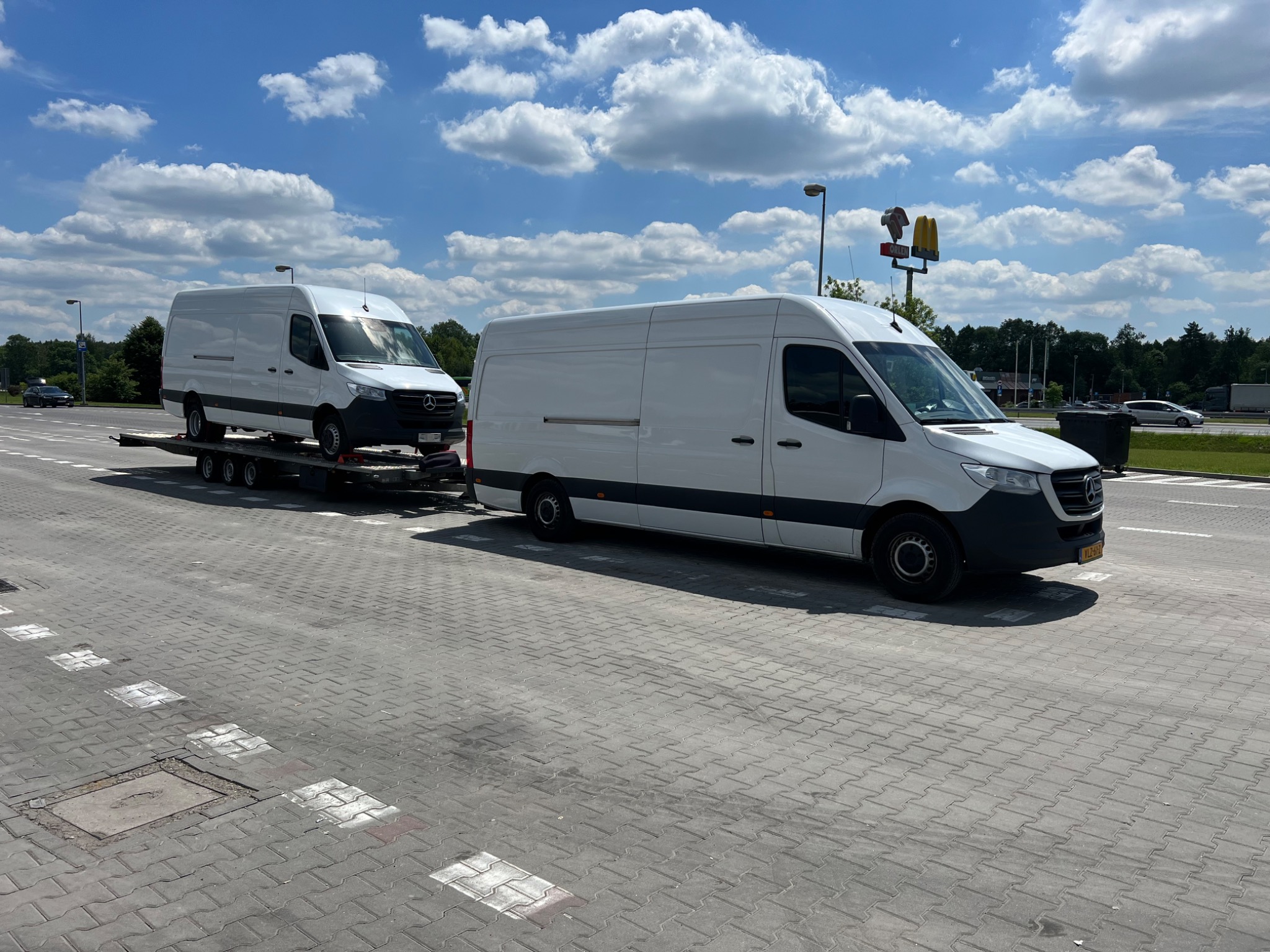 Dwa białe busy Mercedes-Benz Sprinter, jeden na lawecie za drugim, na parkingu w słoneczny dzień. W tle widoczny znak stacji Orlen i McDonald's.